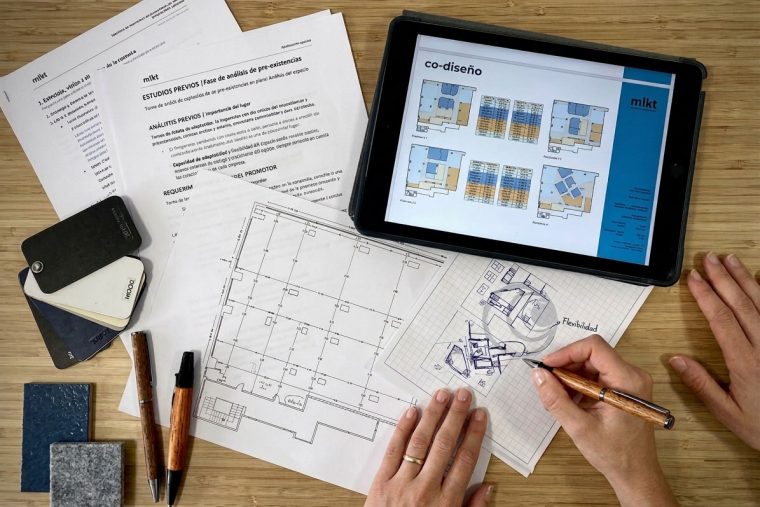 Vista cenital de arquitectos del estudio MLKT trabajando sobre planos de oficinas, bocetos de distribución y muestras de materiales. Representa el proceso estratégico de co-diseño colaborativo junto a promotores para crear espacios de trabajo eficientes, flexibles y adaptados.